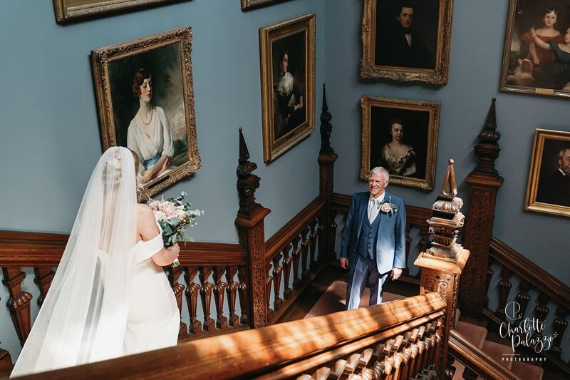Bride's father wearing a blue suit waiting for his daughter, the bride, wearing a white dress walking down the stairs. 