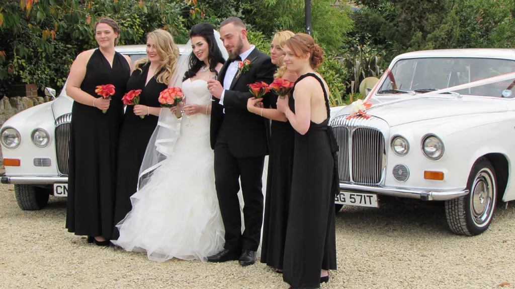 Bride wearing a white dress with her groom wearing a black suit surrounded by all the Bridesmaids wearing matching black dresses and holding an orange themed bouquet of flowers in their hands. Two Classic Cars in the background
