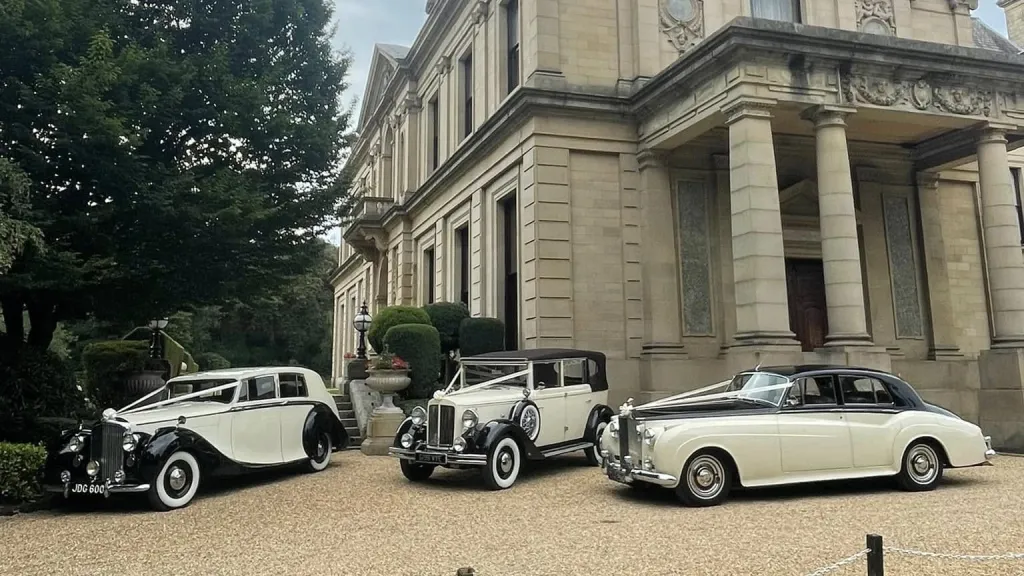 Three matching vintage cars in Black and Ivory with matching white ribbons and bows parked in frotn of a wedding venue