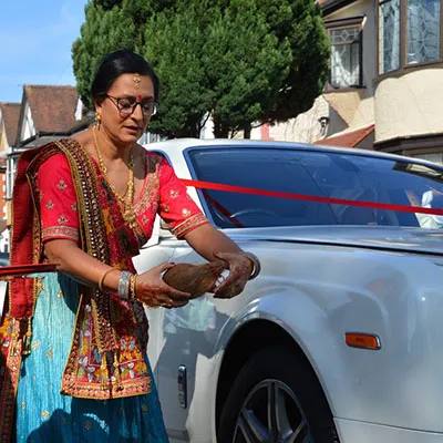 The mother of the Bride, in traditional Indian attire, performing a wedding ritual by placing a coconut in front of the wheel of a white luxury Rolls-Royce car decorated with a red ribbon. She is wearing a red and turquoise lehenga with gold embroidery