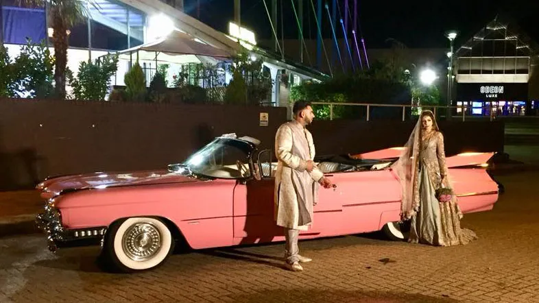 A bride and groom in traditional South Asian wedding attire standing in front of a classic pink American Cadillac convertible at night. The scene is well-lit, with the bride holding a bouquet of flowers and the groom reaching out toward her. The car has large whitewall tires. In the background, modern buildings and neon lights give an urban nighttime ambiance.