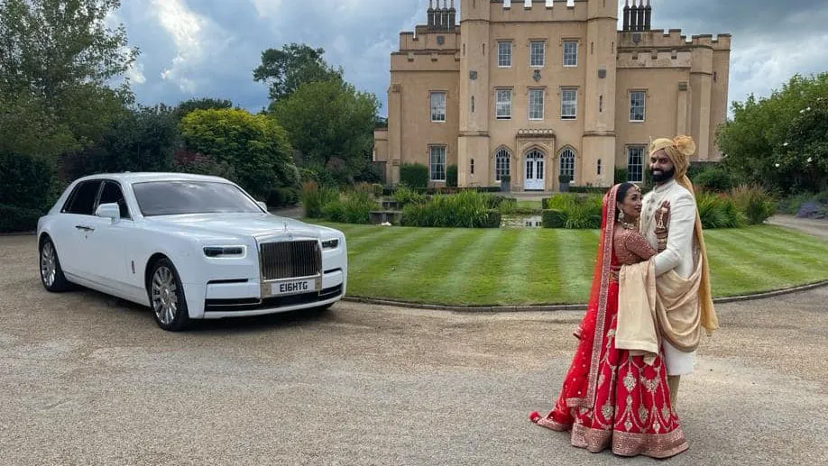 A newly married Asian couple, the bride is wearing a red lehenga and the groom in a cream sherwani. Both are standing holding each others in front of a white Rolls-Royce Phantom, with a castle-like venue behind them