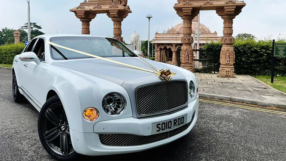 A white Bentley Mulsanne with a silver bonnet decorated with gold ribbons and a bow, parked outside an ornate stone Hindu temple with detailed carvings