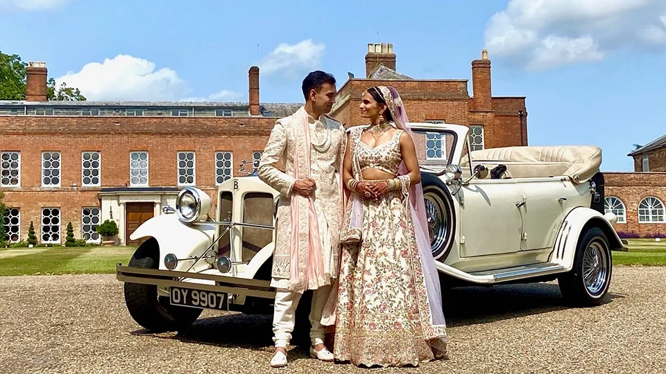 A newly married couple, dressed in traditional Asian light-colored wedding outfits, standing in front of a cream-coloured vintage convertible Beauford car. A large historic manor house is visible behind them.