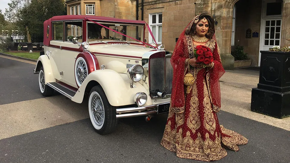 Asian bride in a red and gold traditional wedding gown with a matching veil and gold jewelry, standing beside a cream and burgundy vintage style limousine with wire wheels and a soft top.