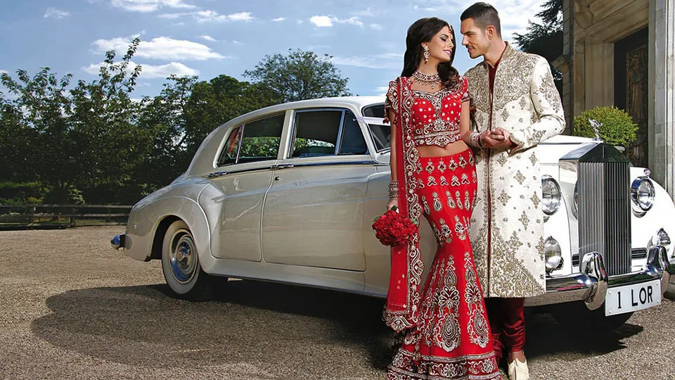 A bride in a red and gold wedding outfit and a groom in a cream and gold sherwani, looking at each other while standing next to a white classic Rolls-Royce Silver Cloud.
