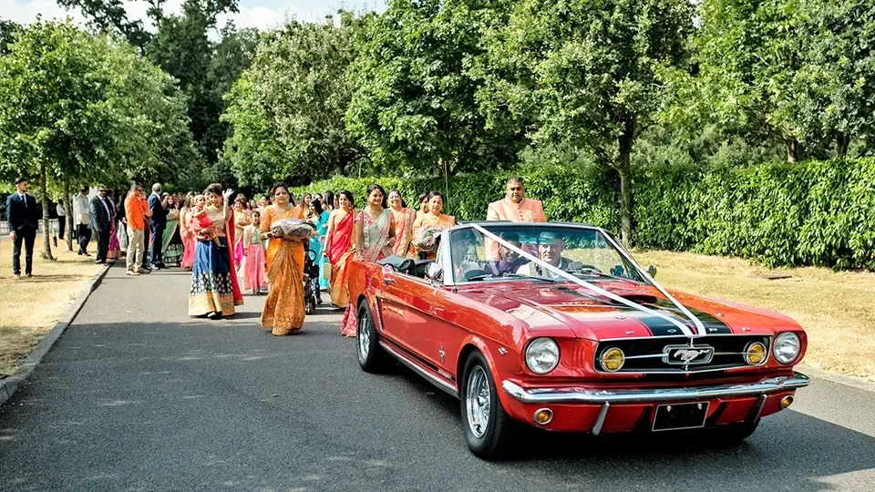 Classic red Ford Mustang convertible decorated with white ribbons, carrying the groom as part of a the Baraat procession followed by guests down a tree-lined path.
