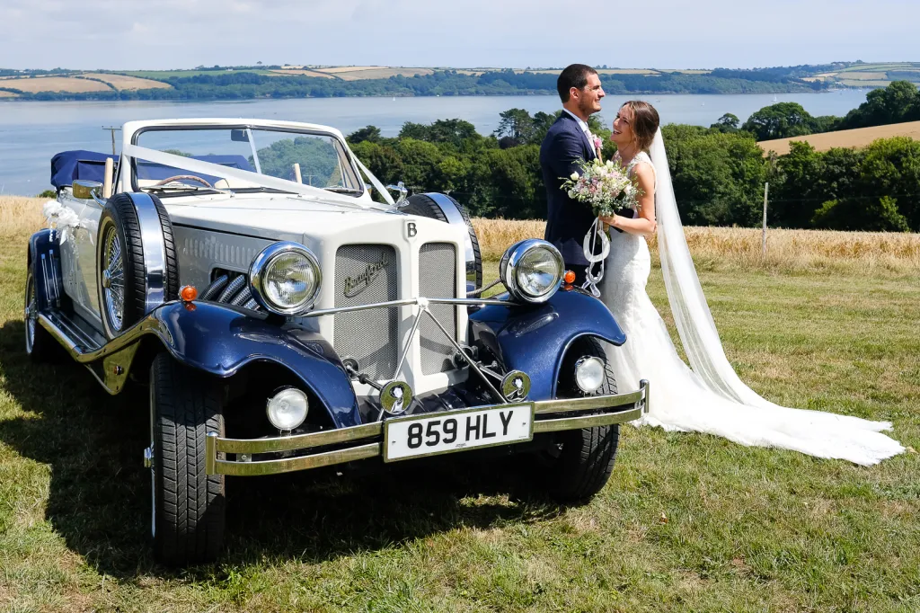 Bride and groom beside a white and navy Beauford convertible wedding car overlooking a lake