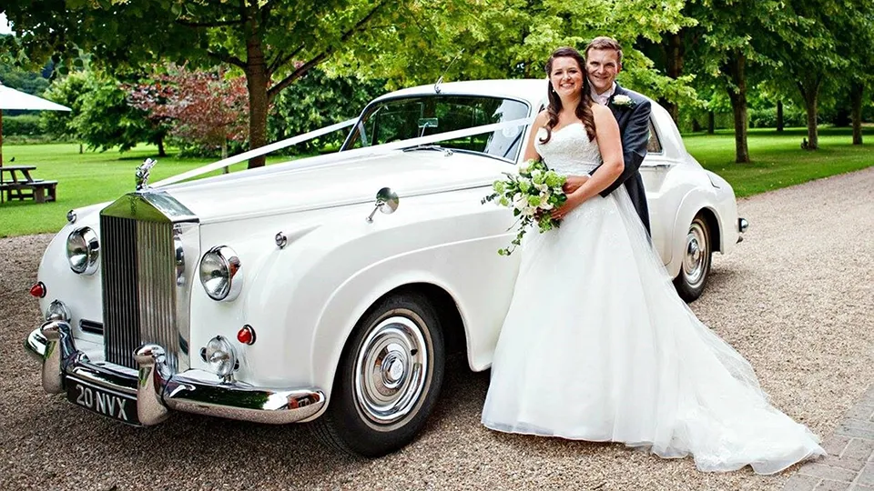 Bride and groom standing by a white Rolls-Royce Silver Cloud with wedding ribbon in a tree‑lined park.