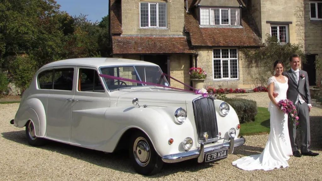 A popular white austin Princess Limousine decorated with purple ribbons matching the Bride's bouquet of flowers she is holding in her hands. Bride and Groom are standing in front iof the vehicle smiling. 