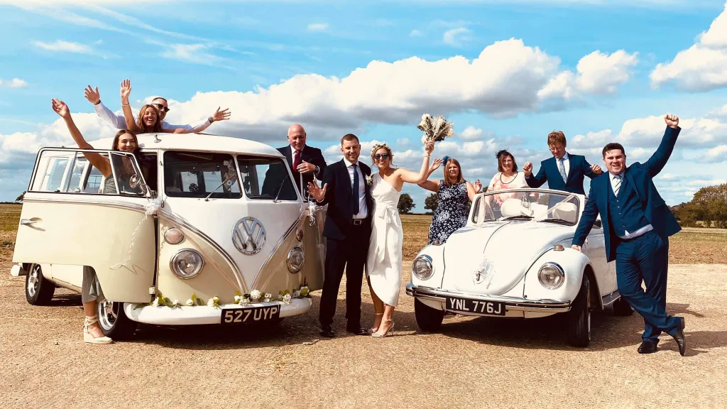 Wedding party cheering between a cream VW Campervan and a white convertible Beetle.