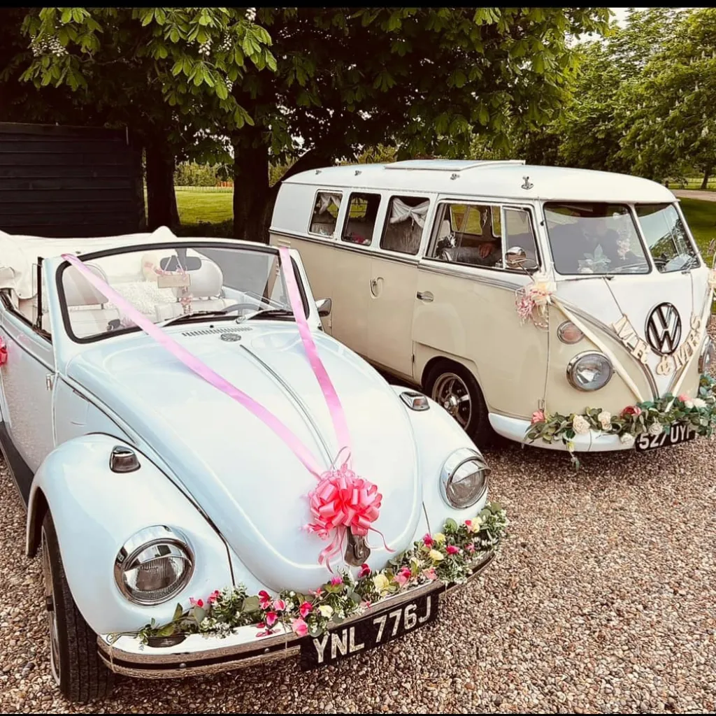 A classic VW Campervan on the right parked next to a classic convertible Beetle on the left dressed with matching and colourful floral arrangement on their front bumper