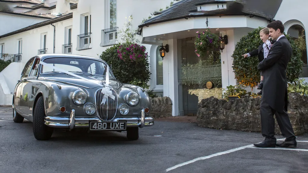 Silver grey classic jaguar Mk2 dressed with white ribbons and bow with groom holding his son in his arm.