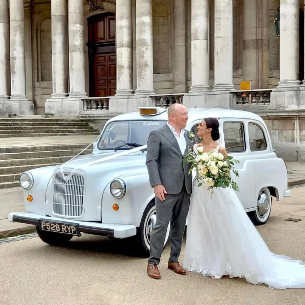 Bride and Groom standing in front of a white classic Taxicab dressed with wedding ribbons