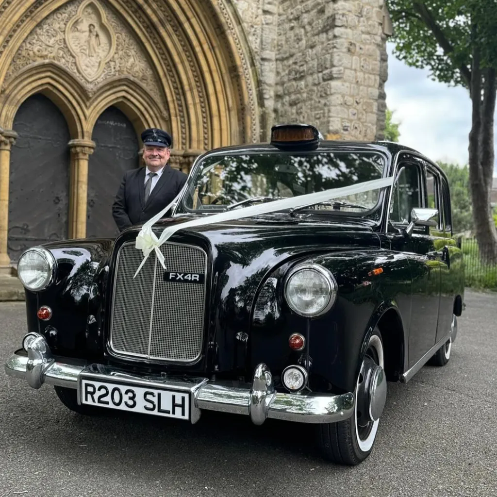 Classic black taxi cab decorated with white ribbons on its front bonnet with fully uniformed chauffeur smiling in the background