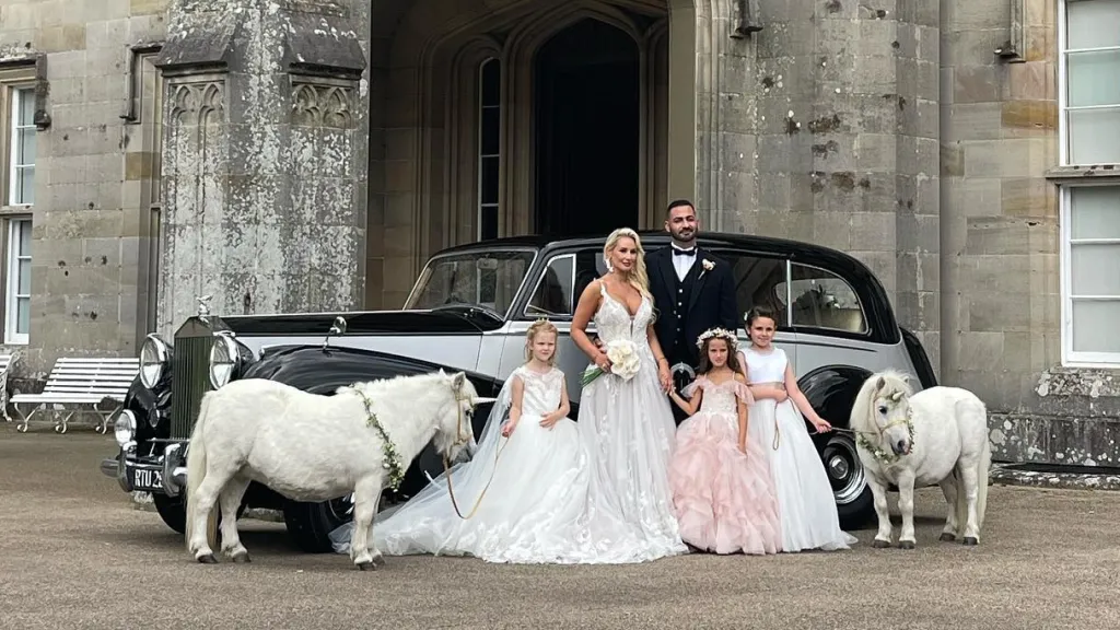 Bride and groom with three flower girls and two white ponies in front of a vintage black and silver Rolls-Royce wedding car outside a grand country house.