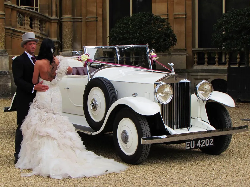Elegant couple beside a white vintage Rolls-Royce convertible decorated with wedding ribbons outside a manor house.