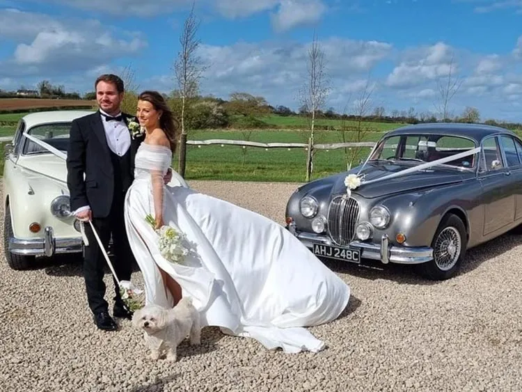 Bride and groom standing with two classic wedding cars, a silver Jaguar and ivory Daimler, on a country estate drive.
