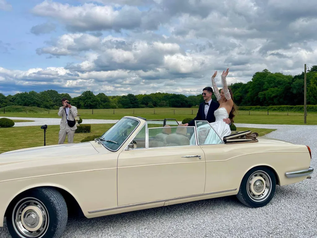 Bride and groom celebrating in a cream Rolls-Royce convertible with the roof down on their wedding day.