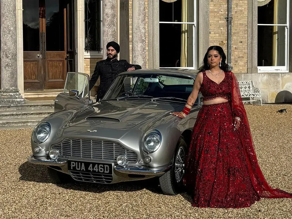 Stylish Asian couple posing beside a silver Aston Martin DB6 outside a grand wedding venue.