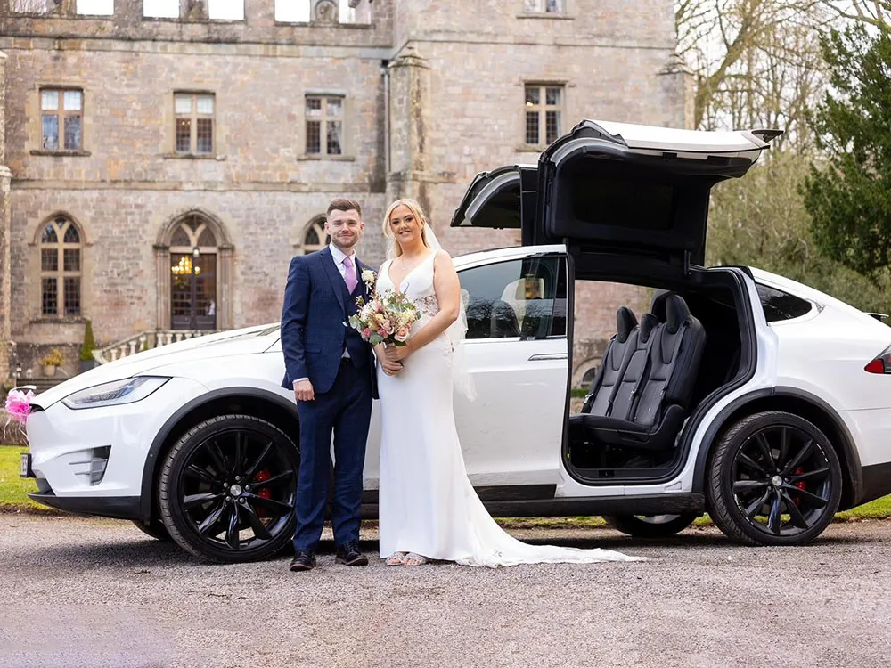 Bride and groom standing beside a white Tesla Model X wedding car with its signature falcon-wing doors open.