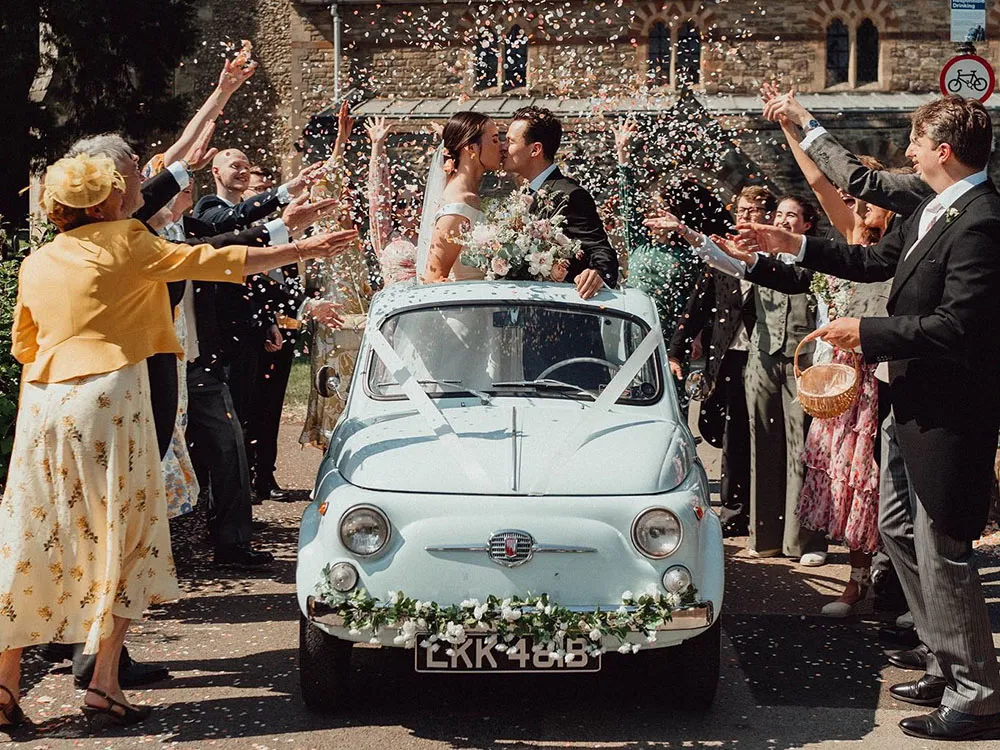 Guests shower the newlyweds with confetti as they sit in a light blue Fiat 500 wedding car decorated with flowers.