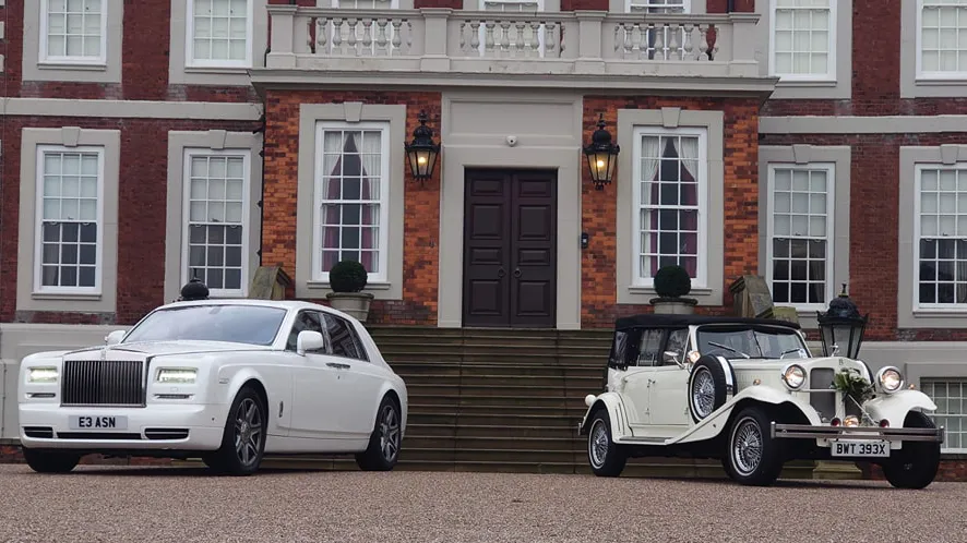 a white Modern rolls-Royce phanto mon the left and a classic vintage white Beauford on the right. Both parked ion frotn of a grand wedding venue