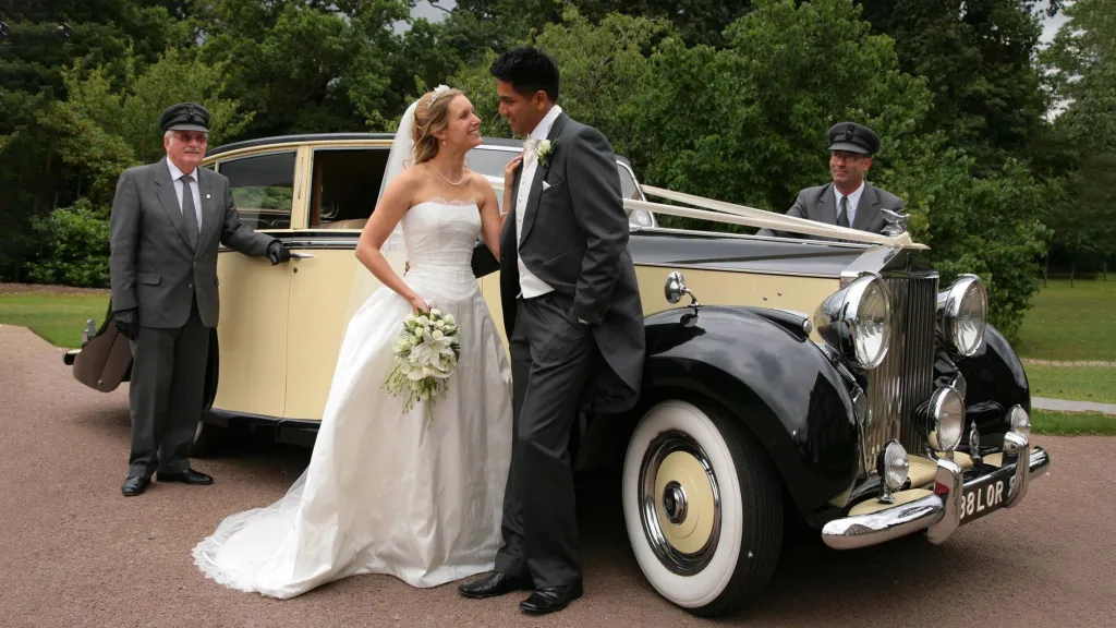 smiling Bride with her groom standing in front of a Black and Ivory vintage Rolls-Royce with two chauffeurs standing by the vehicle wearing a full grey uniform with caps