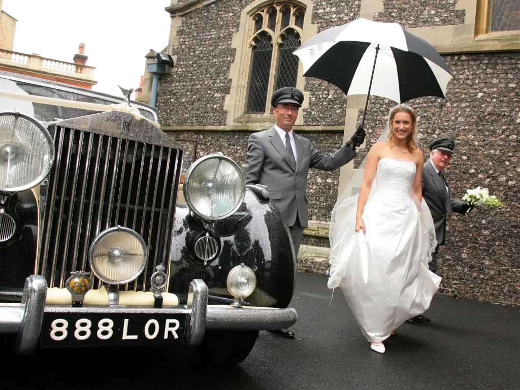 Fully uniformed chauffeur holding a Black & White umbrella over a Bride wearing a white dress. Vintage car in Black & Ivory in the background