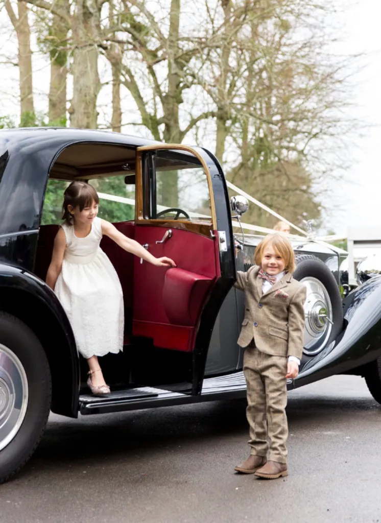Little page boy keeping the door of a vintage car open for the page girl to exit with a white dress