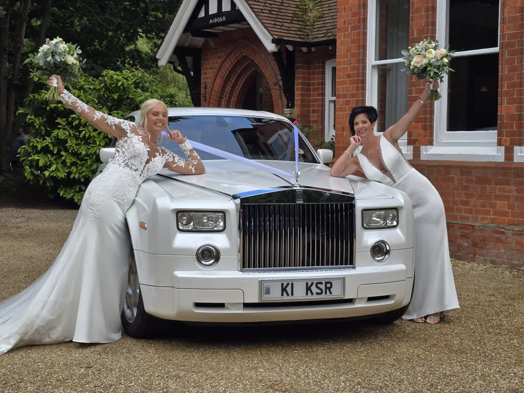 Two smiling brides wearing a white dress leaning on the front bonnet of a modern white Rolls-Royce Phantom 