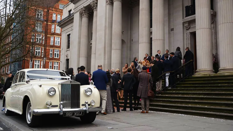 Ivory Classic Rolls-Royce silver Cloud parked outside the main entrance of the Old Marylebone Town Hall. Wedding guests are standing on the stairs leading up to the front entrance door. 