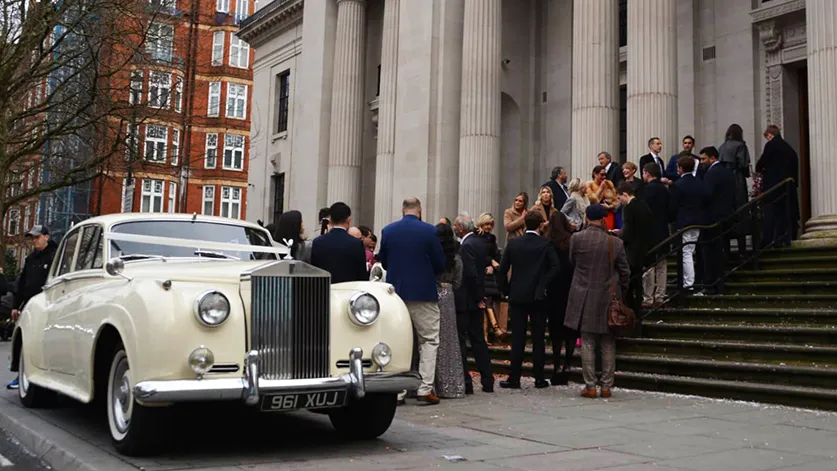 Cream classic Rolls-Royce Silver Cloud wedding car parked outside Old Marylebone Town Hall with wedding guests in background