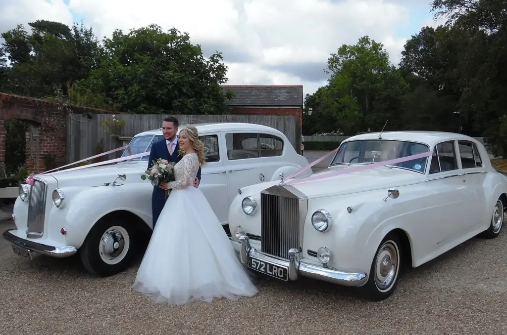 Two classic white wedding cars decorated with traditional V-shaped bonnet ribbons