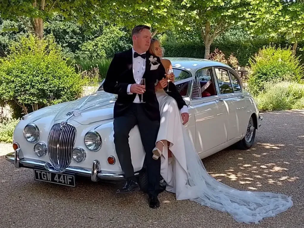 Bride and groom holding champagne, sitting on the front wing of a white Jaguar MK2 at a wedding venue