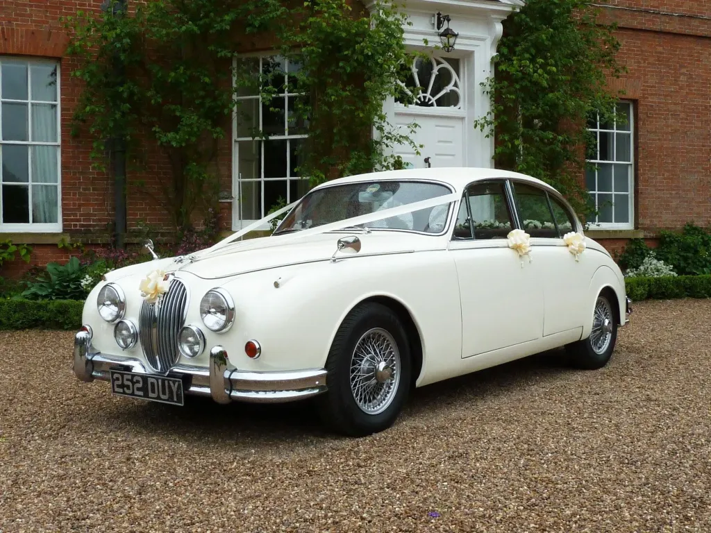 White classic Jaguar MK2 wedding car decorated with ivory ribbons and bows outside a red-brick country house