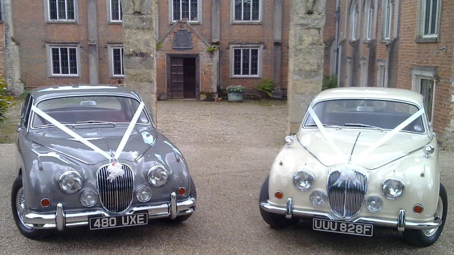 Silver grey Jaguar MK2 and ivory Daimler 250 V8 parked side by side with white wedding ribbons in front of a red-brick manor house