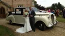 Bride and groom standing in front of an Austin Princess Limousine with semi-convertible roof open. Wedding guests can be seen in the background