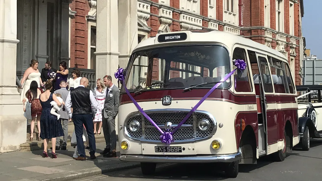 Classic single decker bus decorated with purple ribbons and bows parked in front of registry office with wedding guests in the background