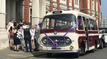 Classic single decker bus decorated with purple ribbons and bows parked in front of registry office with wedding guests in the background
