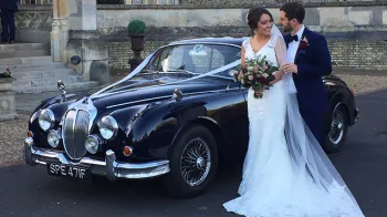 Groom holding bride in his arms with Dark blue Daimler 250 V8 in the background