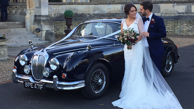 Groom holding bride in his arms with Dark blue Daimler 250 V8 in the background