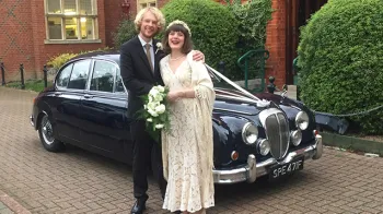 Groom holding bride in his arms with Dark blue Daimler 250 V8 in the background