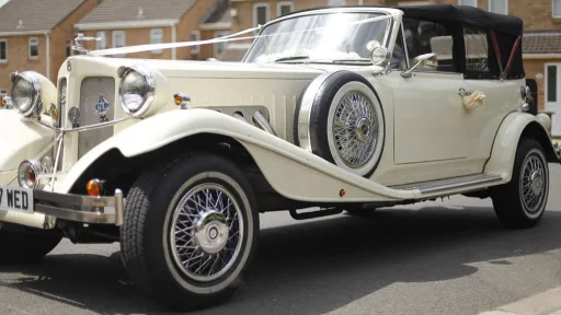 Front left side view of Vintage Beauford decorated with white ribbbon, spare wheel mounted on the side of the vehicle with thin white wall tires.