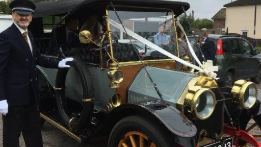 Close up view of Vintage Oakland with its chauffeur standing by the vehicle wearing s full suit with hat and white gloves