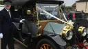 Close up view of Vintage Oakland with its chauffeur standing by the vehicle wearing s full suit with hat and white gloves
