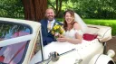 Bride and groom smiling seating inside convertible Beauford Convertible. Bride is holding a bouquet of uyellow flowers in her hands.