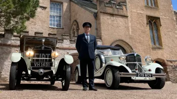 Fully uniformed chauffeur standing in the middle of two vintage cars. Car on the right is a convertible Beauford and the left car a Vintage Fiat