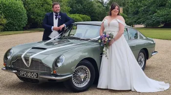 Aston Martin decorated with ivory ribbons with Bride and Groom standing on either side of the vehicle