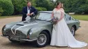 Aston Martin decorated with ivory ribbons with Bride and Groom standing on either side of the vehicle